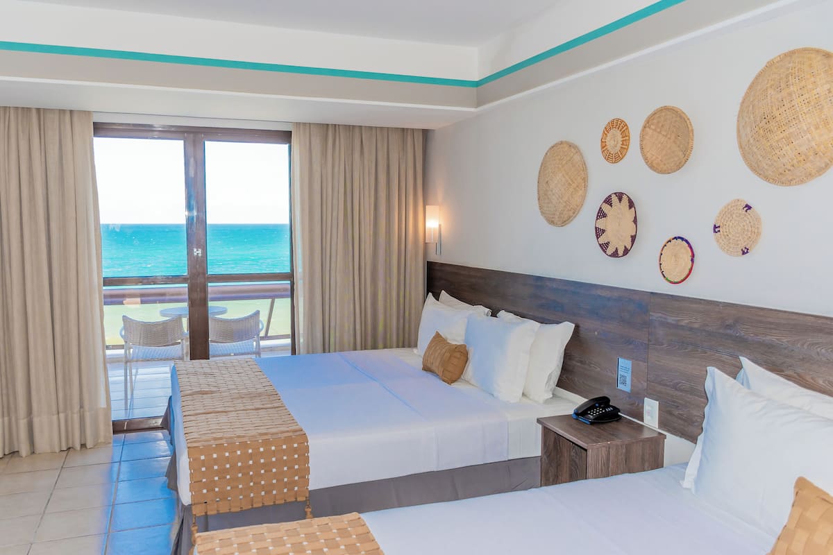 A hotel room with two beds, a balcony with a view of the sea, and decorative woven items on the wall.