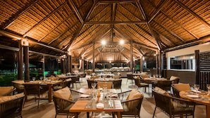 An indoor dining area with multiple tables and chairs, a high ceiling with a thatched roof, and a view of the outdoors.