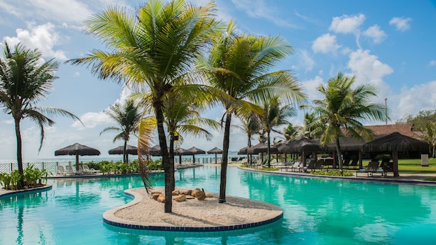 A pool with palm trees and lounge chairs, surrounded by umbrellas and a view of the ocean.