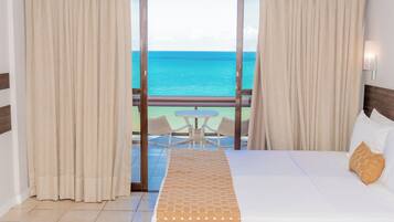 Habitación de hotel con cama, vista al océano, balcón con mesa y sillas, y cortinas.