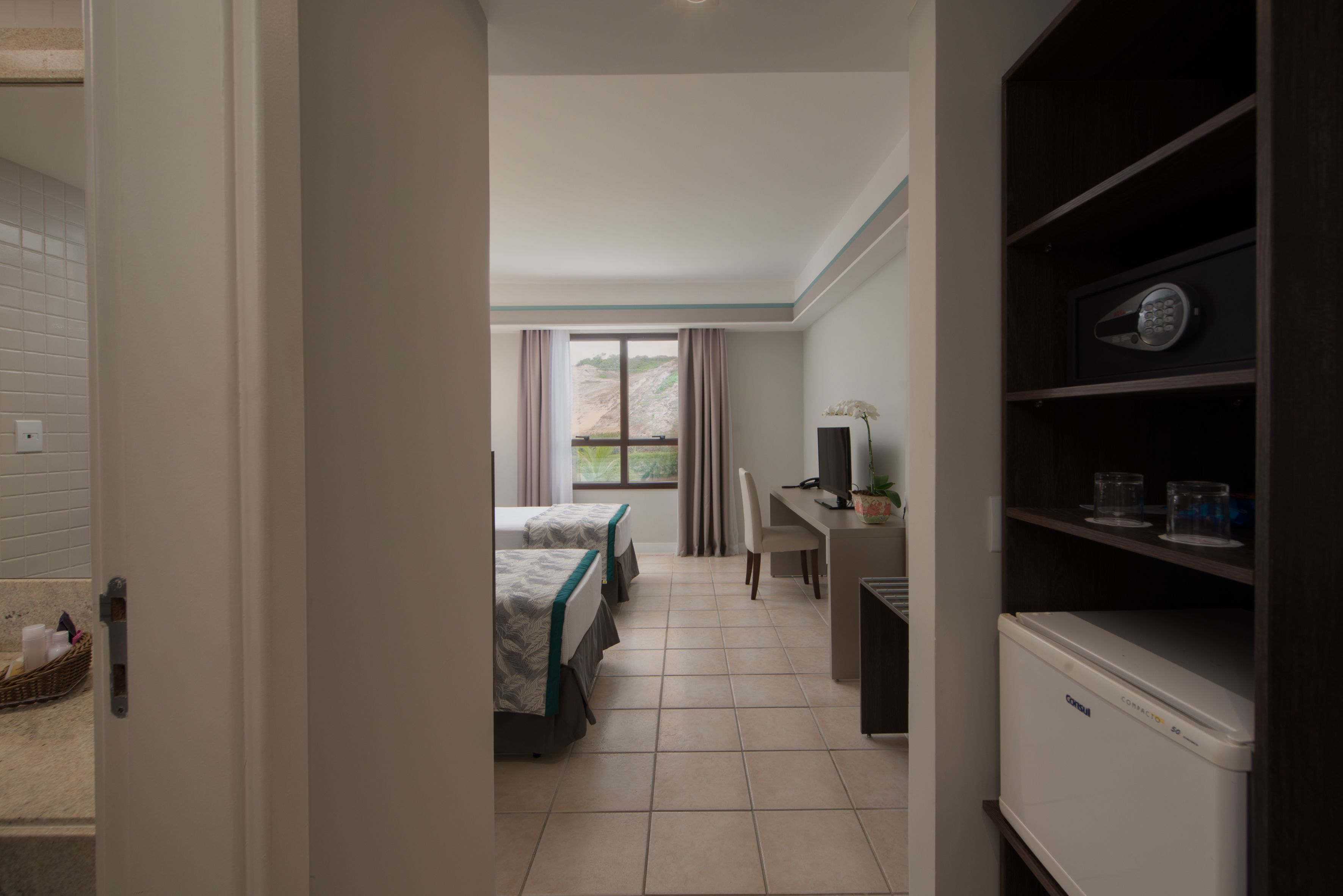 Quarto de hotel com cama, escrivaninha, cadeira, prateleira com objetos e janela com cortinas.