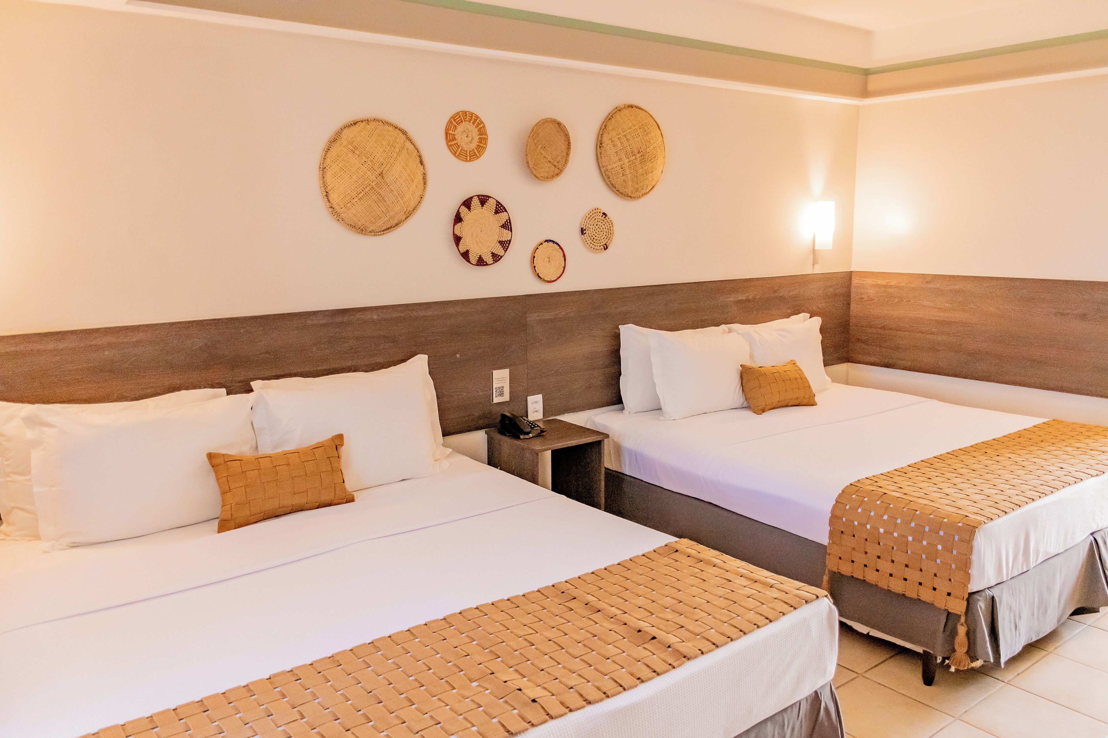 A hotel room with two beds, a wooden headboard, a wall-mounted light, and woven wall art.