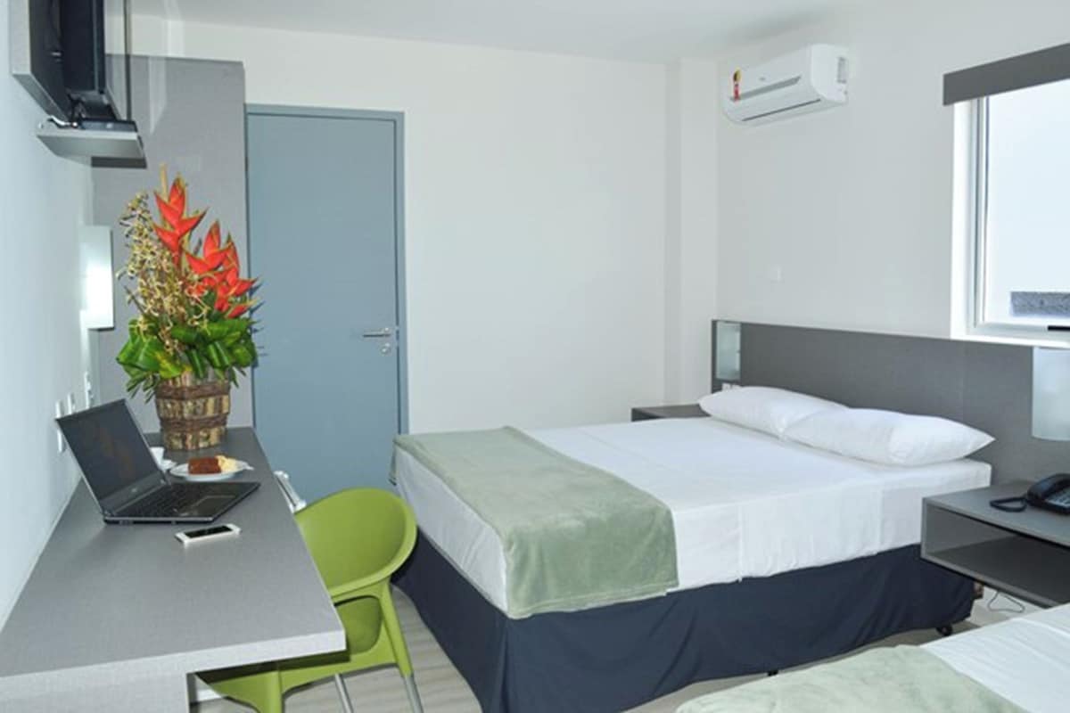 A hotel room with a bed, a desk with a laptop, a chair, and a plant.