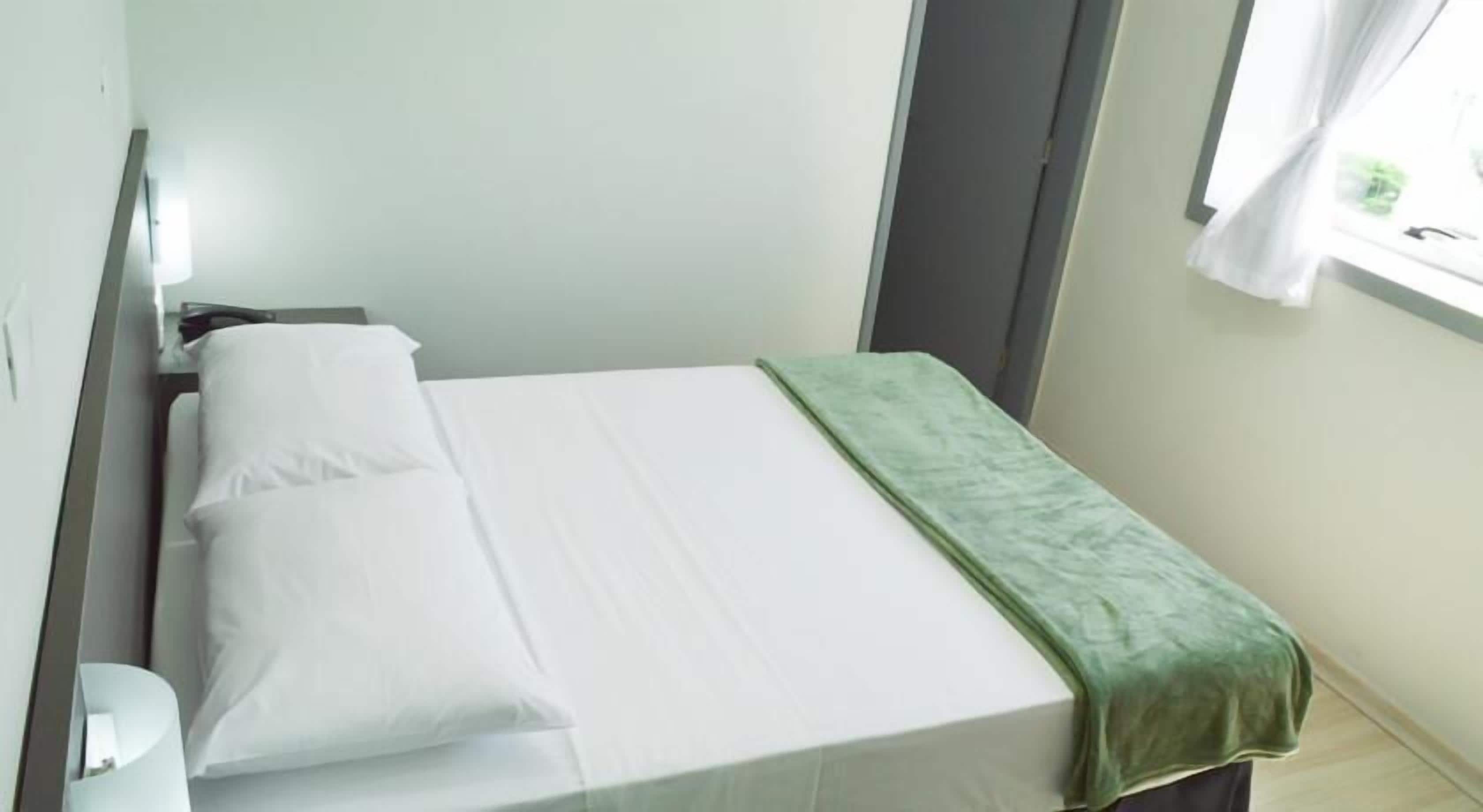 A small, single-bed room with a green bedspread, white pillows, and a window with white curtains.