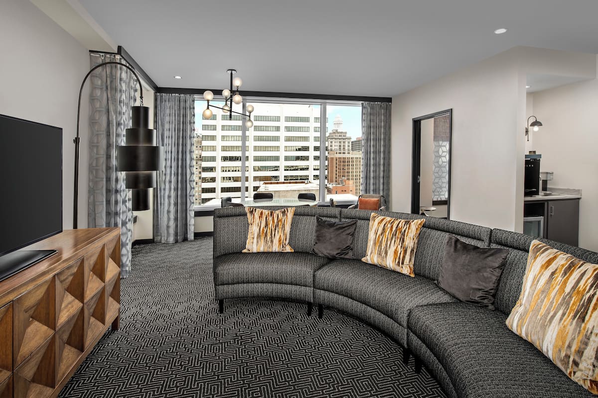 A modern living room with a curved sectional sofa, a wooden TV stand, and a city view through large windows.
