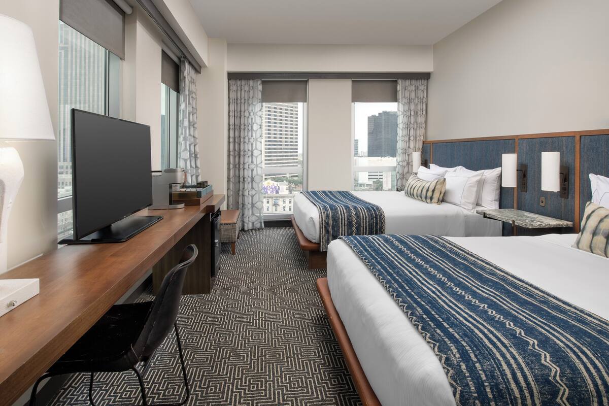 A hotel room with a bed, a desk with a TV, a chair, and a view of the city.