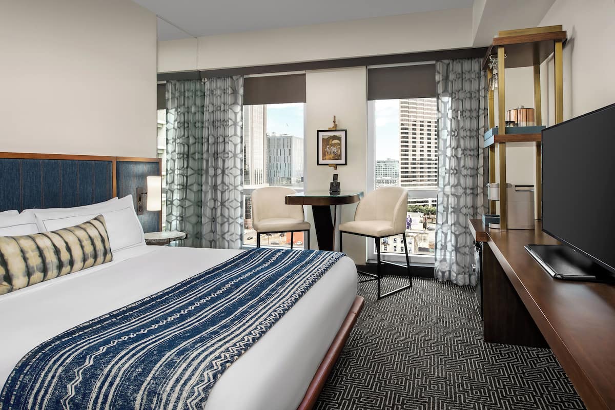 A hotel room with a large bed, two chairs, a small table, a TV, and a city view.