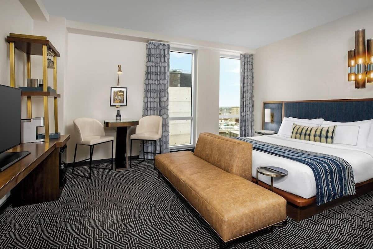 A hotel room with a bed, a desk, a chair, a bench, and a large window.