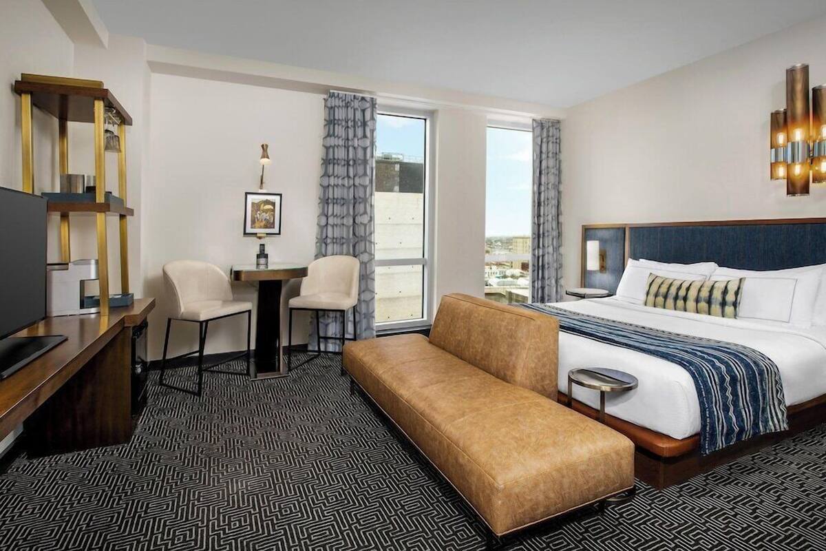 A hotel room with a bed, a desk, a chair, a bench, and a large window.