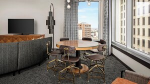 Habitaci ón de hotel moderna con una mesa de comedor redonda, seis sillas, un televisor de pantalla plana y un amplio ventanal con vistas a la ciudad.