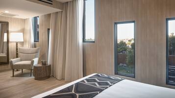 A hotel room with a large bed, a chair, a side table, and a view of the outdoors through large windows.