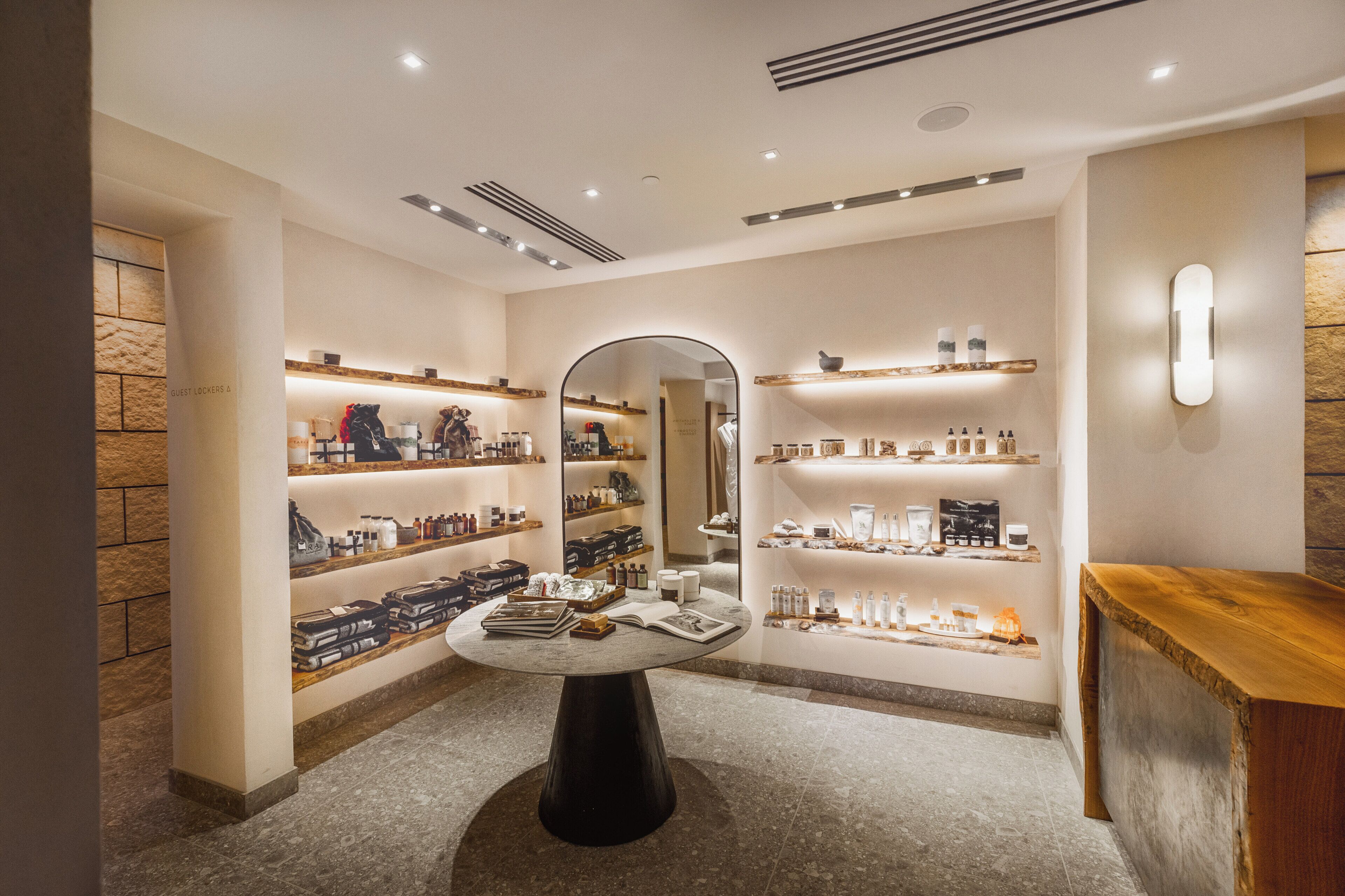 A modern boutique hotel lobby with a central round table, mirrored wall, and shelves displaying various items.