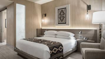 A modern hotel room with a large bed, a bedside table with a lamp, a chair, and a wall-mounted artwork.
