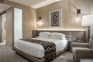 A modern hotel room with a large bed, a bedside table with a lamp, a chair, and a wall-mounted artwork.