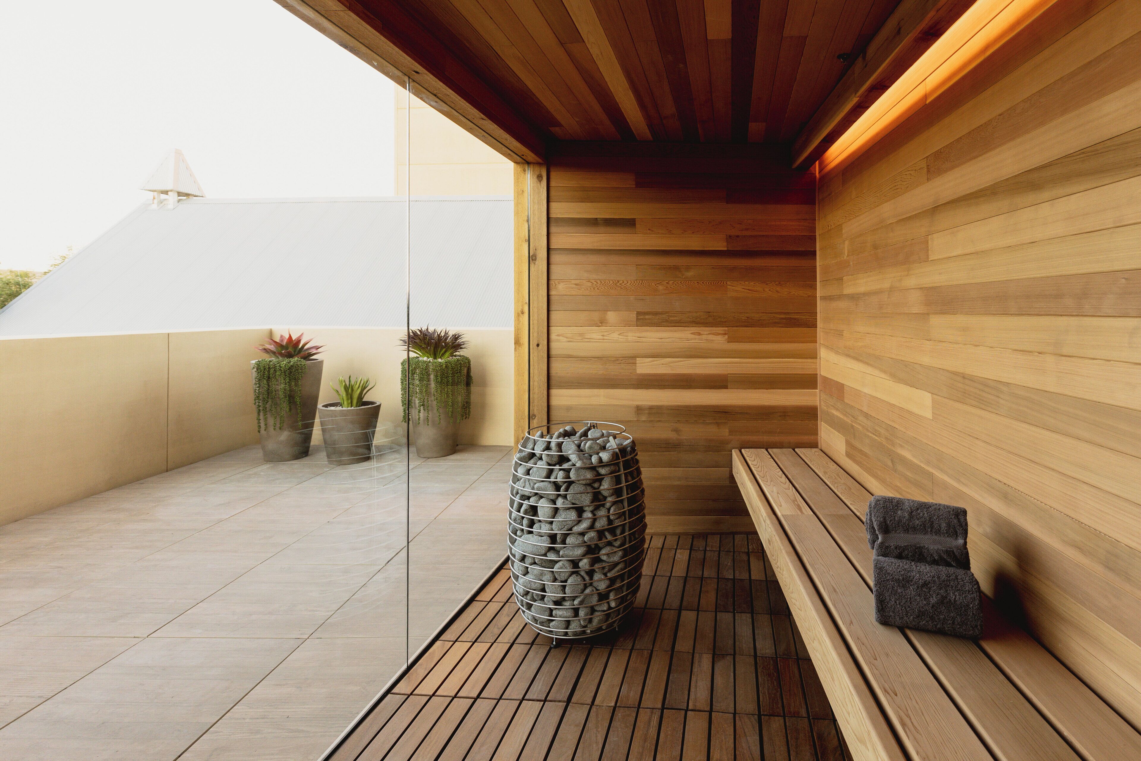 A wooden sauna with a bench and a stone container, potted plants, and a tiled floor.