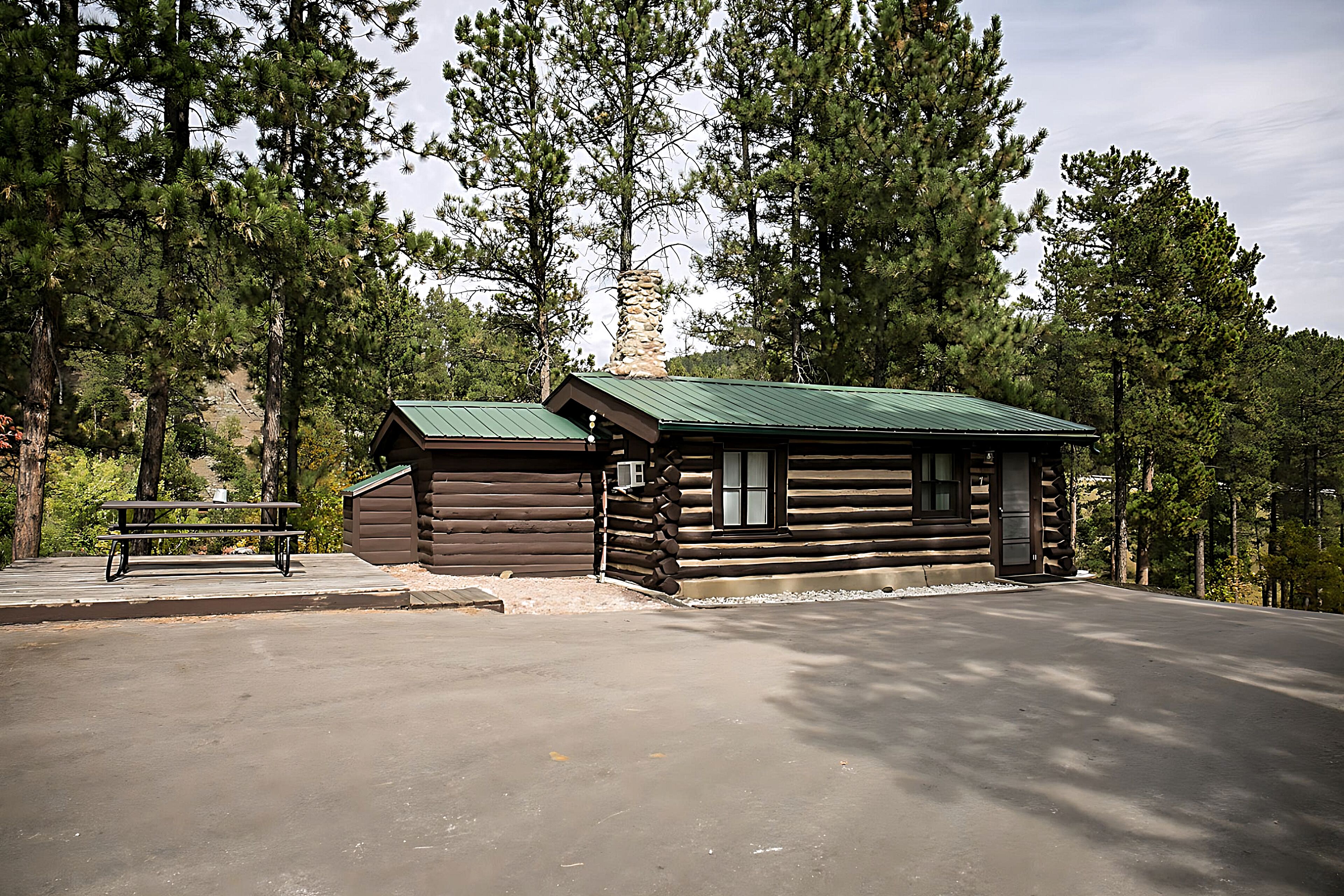 Una cabaña de troncos con techo verde, chimenea de piedra y una mesa de picnic cerca.