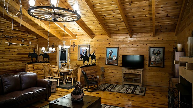 Interior acolhedor de uma cabana de troncos, com um sofá de couro, mesa de jantar de madeira e uma televisão na parede.