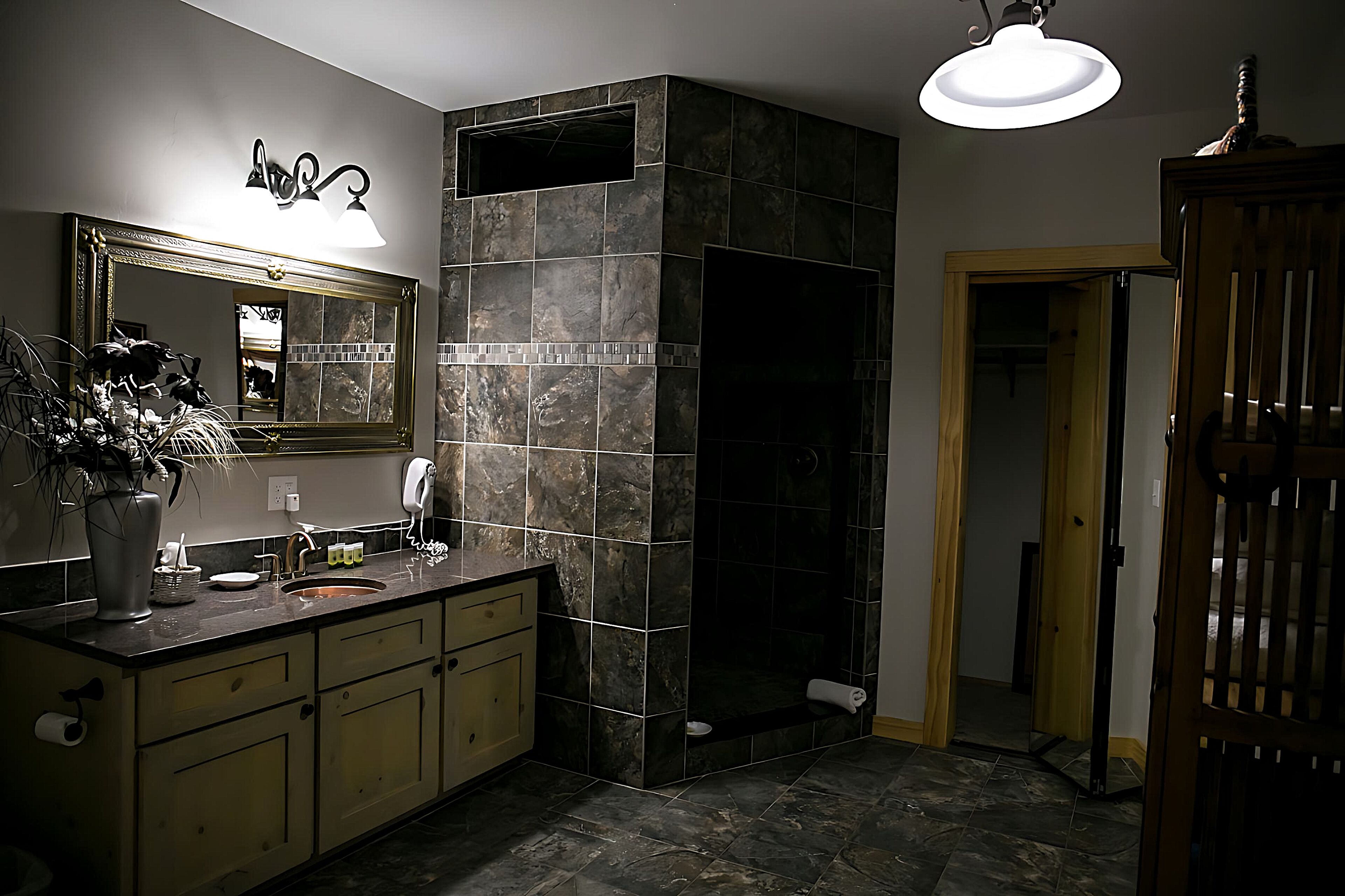 A modern bathroom with a large mirror, dark tiles, a sink, and a shower area.