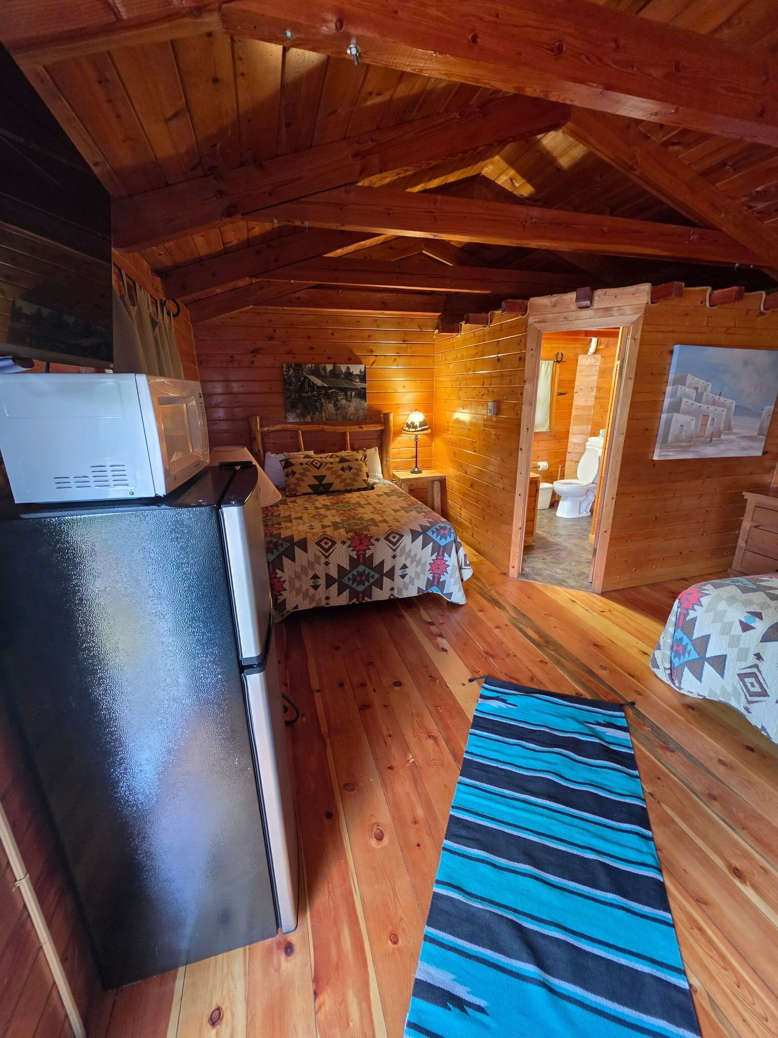 Een knus vakantiehuisje met een kitchenette, een bed met beddengoed met dessin, een blauw gestreept vloerkleed en een schilderij aan de muur.