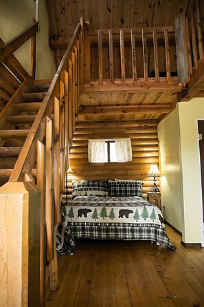 A wooden cabin bedroom with a bed, a nightstand, a lamp, and a wooden staircase.