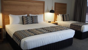 A hotel room with two beds, each with a wooden headboard, white bedding, and patterned throw pillows. There is a nightstand with a lamp between the beds. - Euston Motel (Euston)