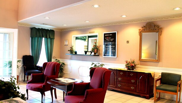 A hotel lobby with a reception desk, two red armchairs, a wooden cabinet, and a large mirror. - American Motel (Lenoir)