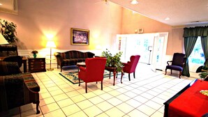 A hotel room with a living area featuring a sofa, chairs, a small table, and a potted plant. There is a door leading to another room and a window with curtains. - American Motel (Lenoir)