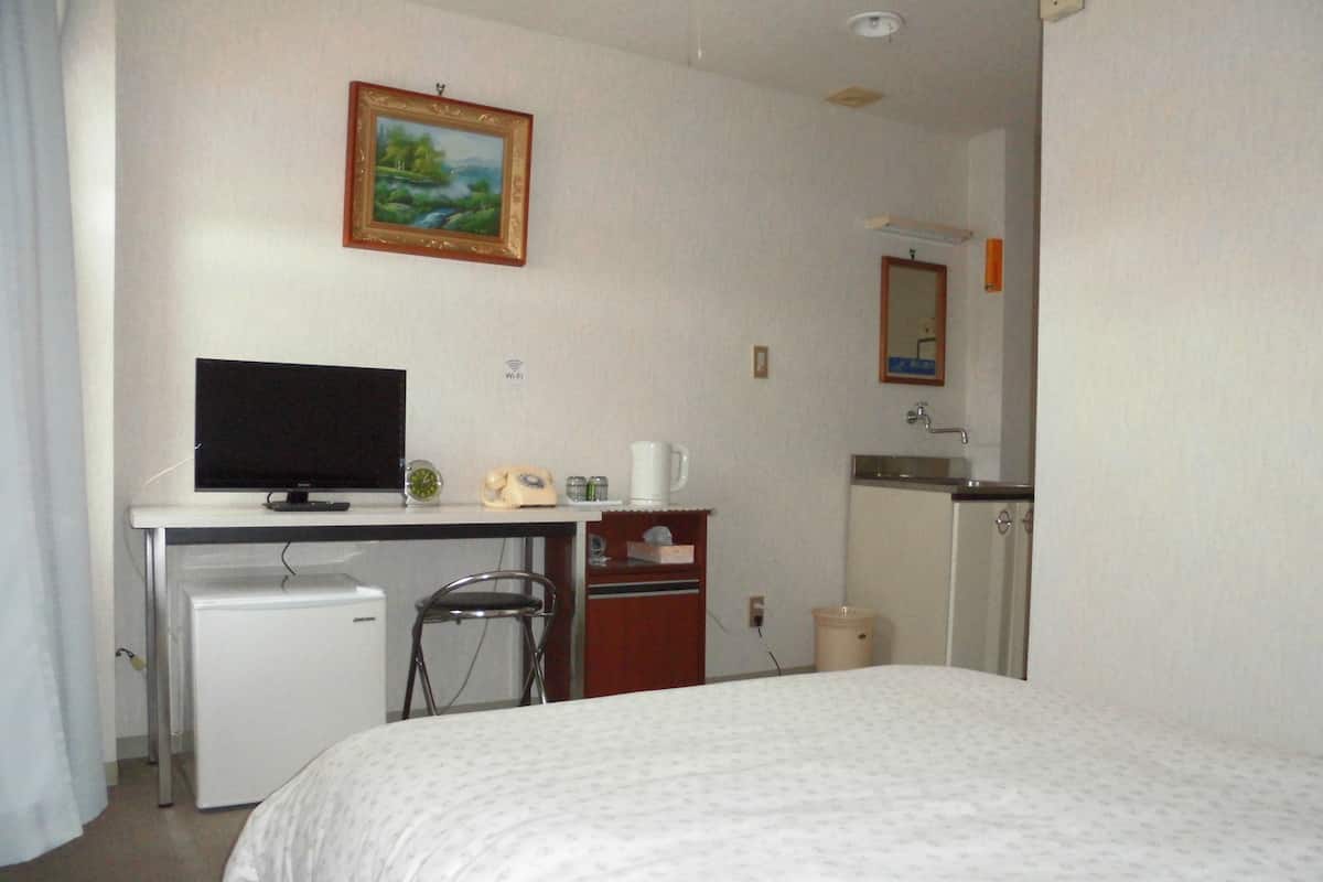 Hotellihuone, jossa on sänky, työpöytä, jossa on televisio, jääkaappi, tuoli, pieni kaappi, peili ja taulu seinällä.