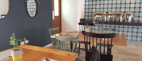 Un comedor con una mesa de madera, sillas y una planta en maceta. Hay dos espejos en la pared y una zona de cocina con salpicadero de azulejos.