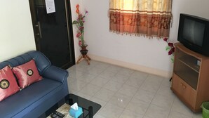 A living room with a blue sofa, a television on a wooden cabinet, a potted plant, and a window with patterned curtains. - Pakasan Garden Home (Khon San)