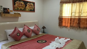 A neatly made bed with a red and beige bedspread, two red pillows with decorative patterns, a framed floral painting above the bed, a wooden shelf with items, and a window with a patterned curtain. - Pakasan Garden Home (Khon San)