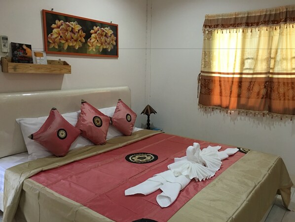 A neatly made bed with a red and beige bedspread, two red pillows with decorative patterns, a framed floral painting above the bed, a wooden shelf with items, and a window with a patterned curtain. - Pakasan Garden Home (Khon San)