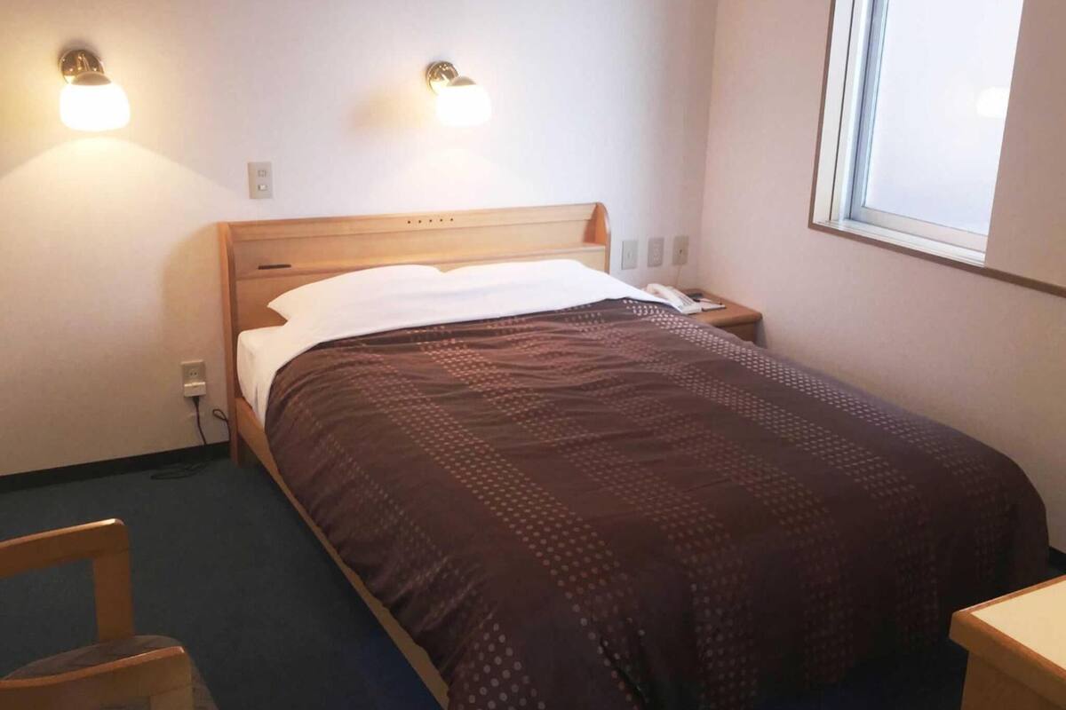 Un lit simple avec une tête de lit en bois, une couvre-lit marron à motifs, deux lampes fixées au mur et une fenêtre sur le côté droit.