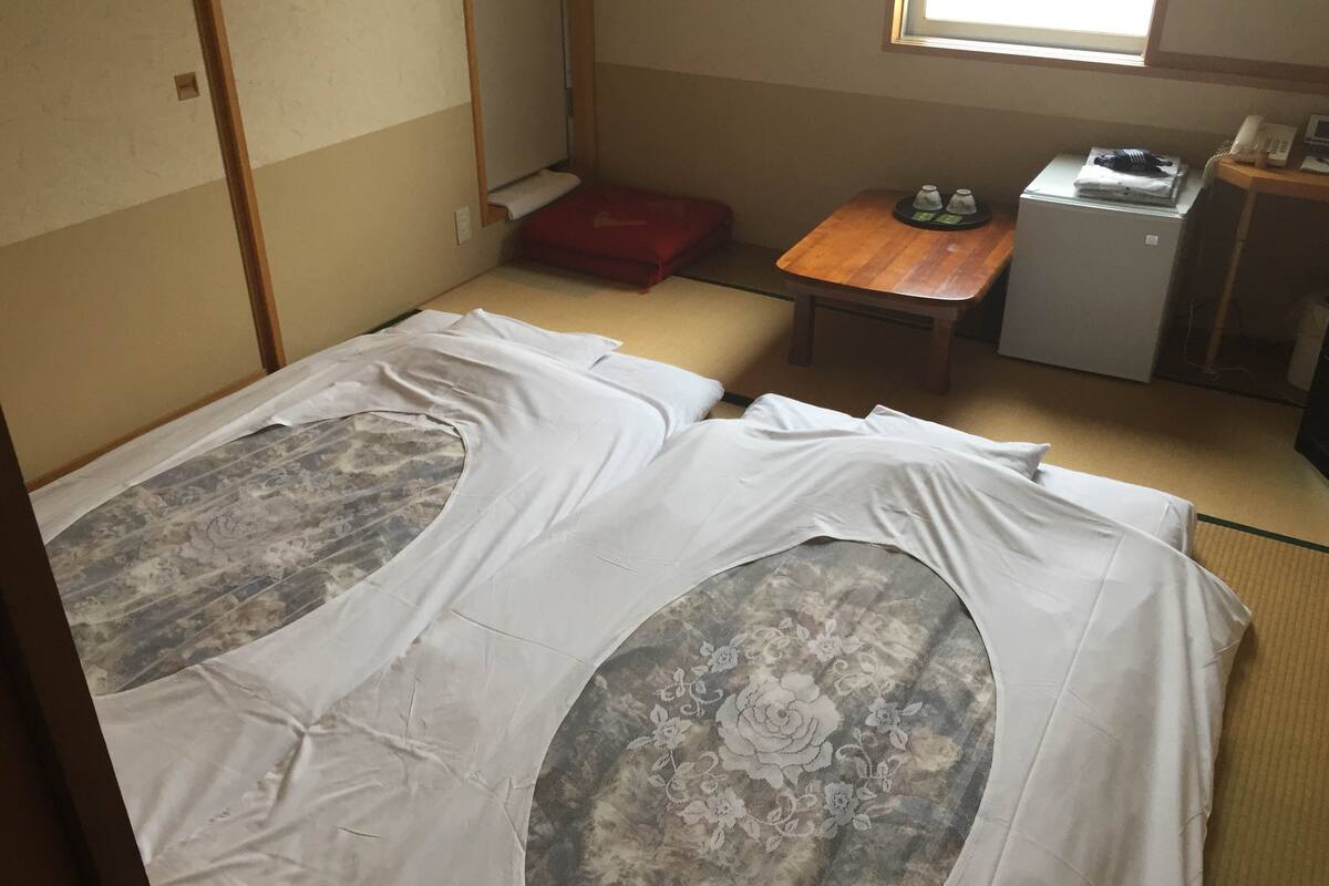 Une pièce de style japonais traditionnel, avec un sol recouvert de tatamis, un futon servant de lit, une petite table en bois et une kitchenette compacte.