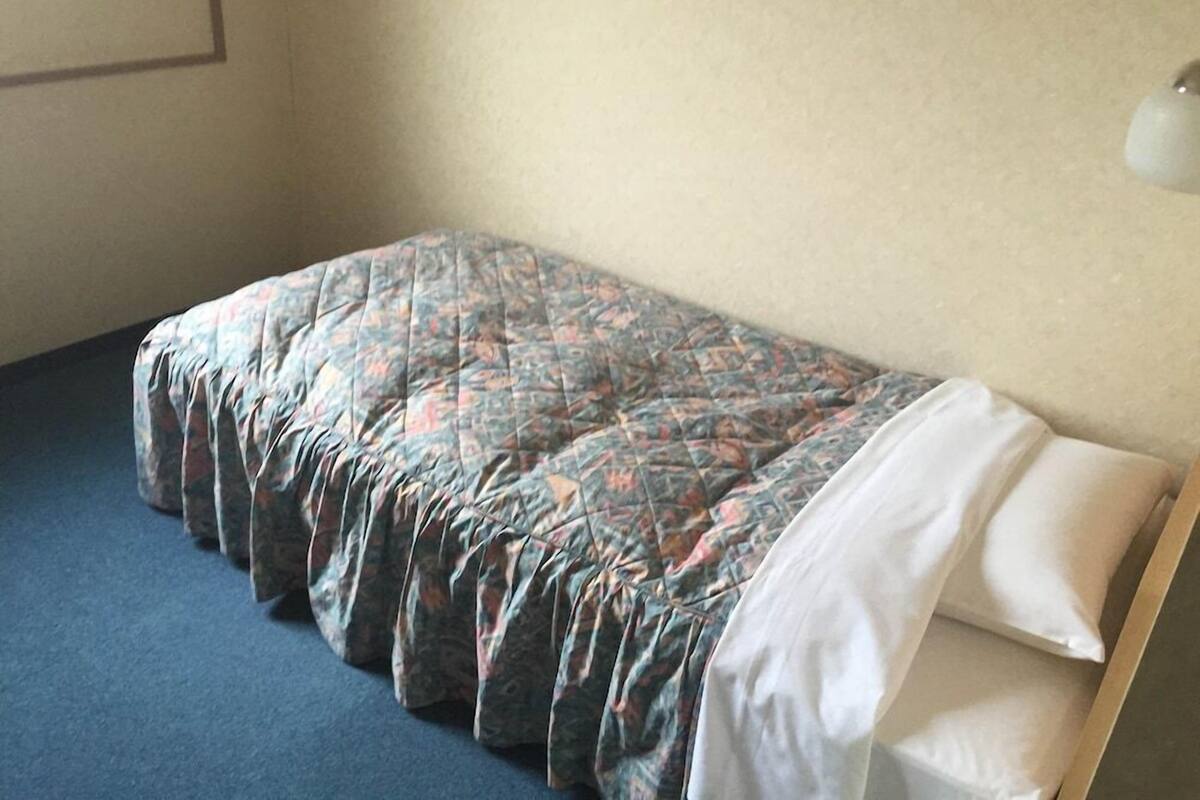 Un lit simple avec une couvre-lit à motifs et des oreillers blancs, dans une pièce dont le mur est uni et qui est éclairée par un luminaire.