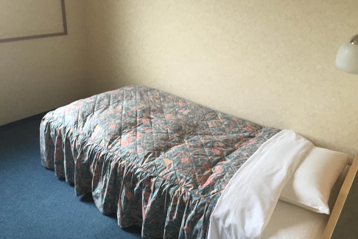 Un lit simple avec une couvre-lit à motifs et des oreillers blancs, dans une pièce dont le mur est uni et le tapis bleu.