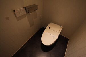 A small, compact bathroom with a toilet, a wall-mounted flush handle, and a trash bin.