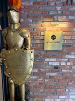 Una estatua de un caballero medieval con escudo, de pie junto a un muro de ladrillo con el letrero 'Soma Hotel'.