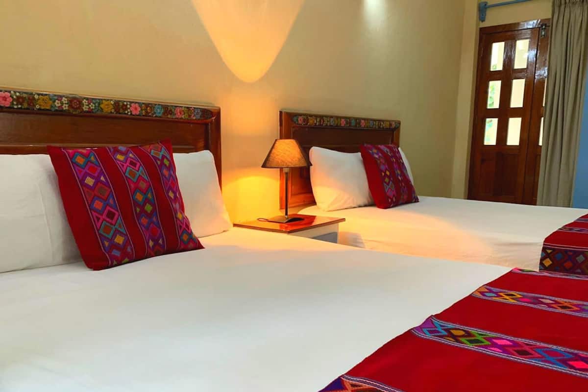 Ein Hotelzimmer mit zwei Betten, jeweils mit weißen Leinentüchern und farbenfrohen gemusterten Kissen. Dazwischen befindet sich ein Nachttisch mit einer Lampe.