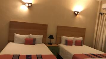 Habitación de hotel con dos camas, cada una con ropa blanca y detalles rojos, una mesita de noche con lámpara, e iluminación en la pared.