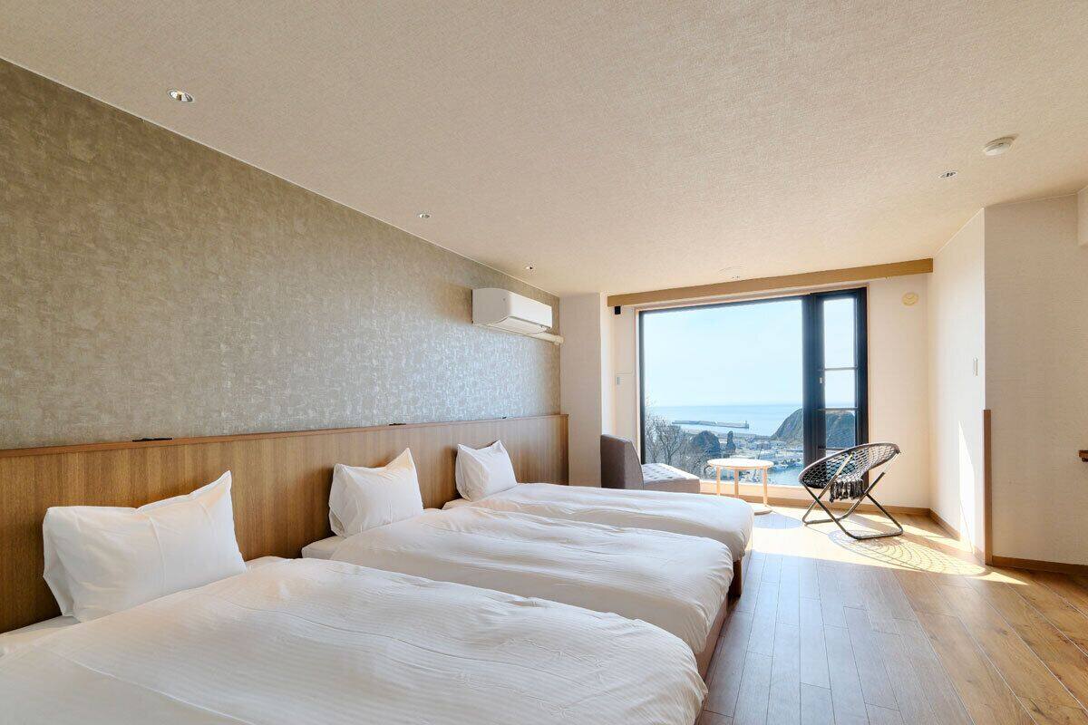 A modern hotel room with three beds, a large window, and a view of the city.