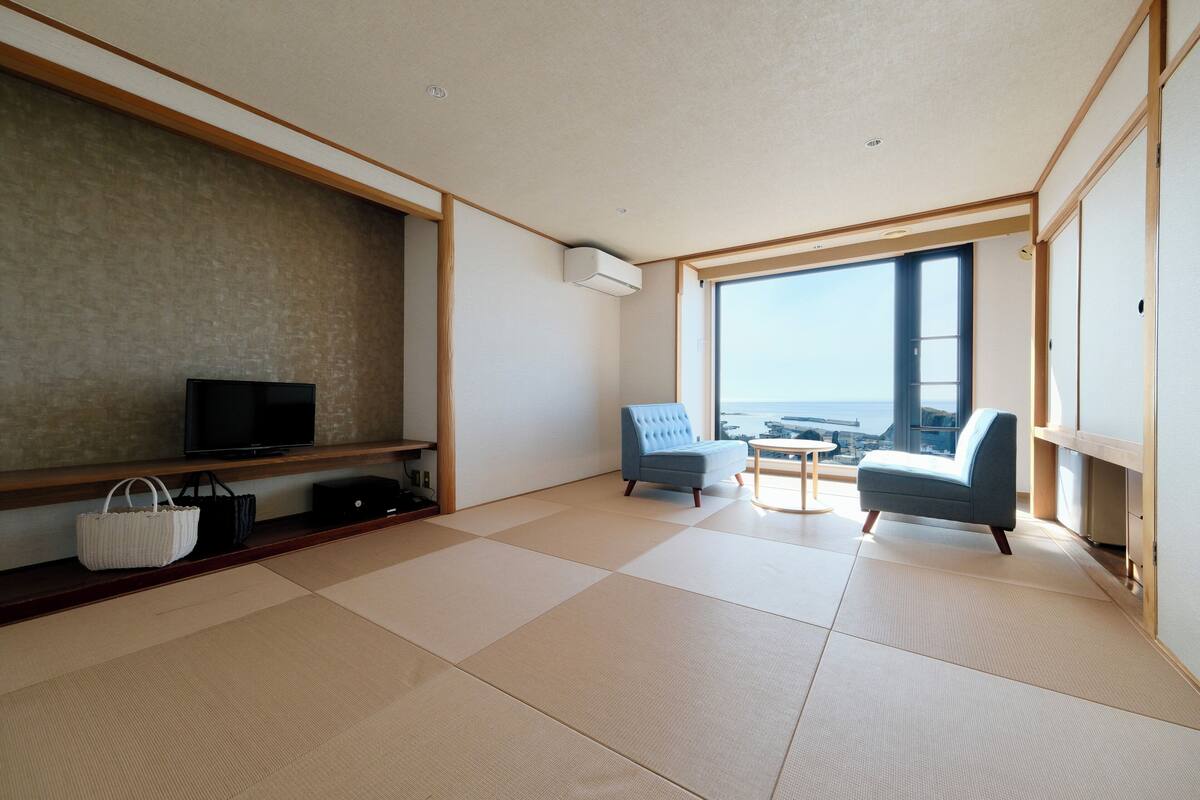 A modern living room with tatami flooring, a flat-screen TV, a sofa, and a view of the sea.