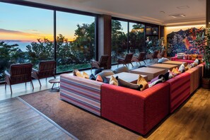 Un salón moderno con un amplio ventanal que ofrece vistas al océano y a los árboles. Hay varias zonas de estar con sofás y sillones.