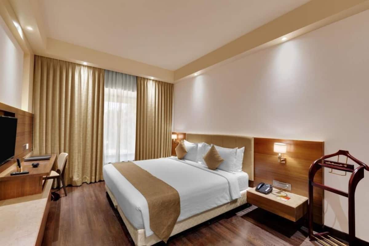 A hotel room with a bed, bedside table, lamp, TV, desk, chair, and curtains.