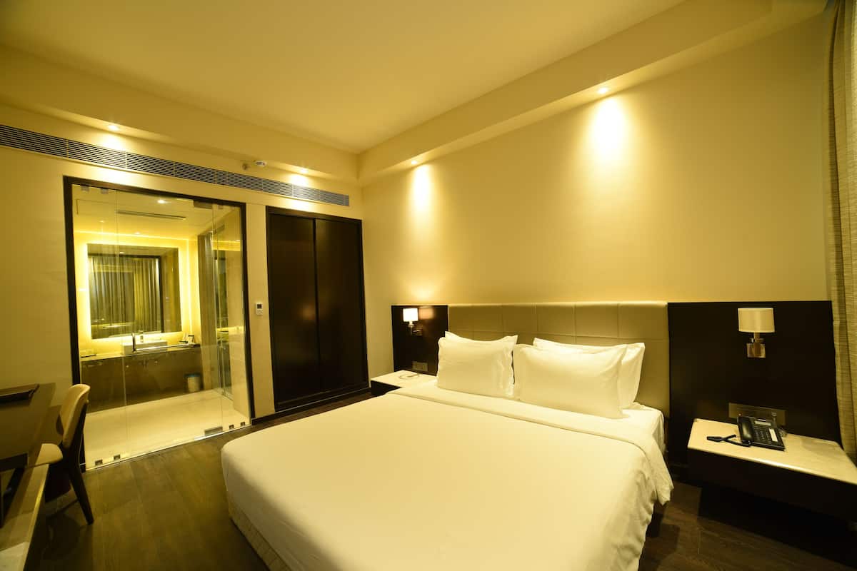 A hotel room with a large bed, a desk, a chair, a nightstand, and a bathroom with a glass shower enclosure.