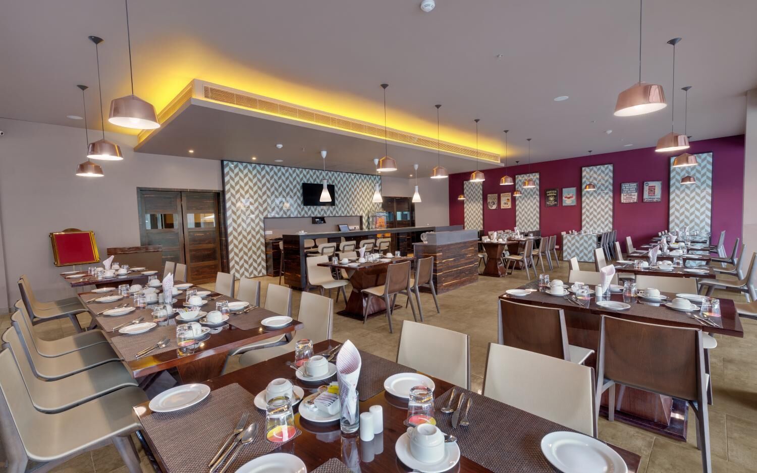 A hotel dining area with multiple tables set for breakfast, a central bar area, and a television mounted on the wall.