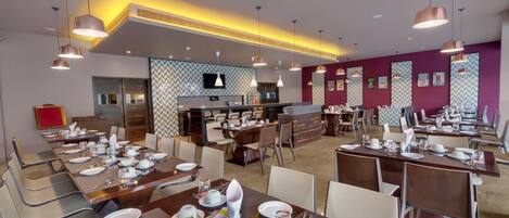 A hotel dining area with multiple tables set for breakfast, a central bar area, and a television mounted on the wall.