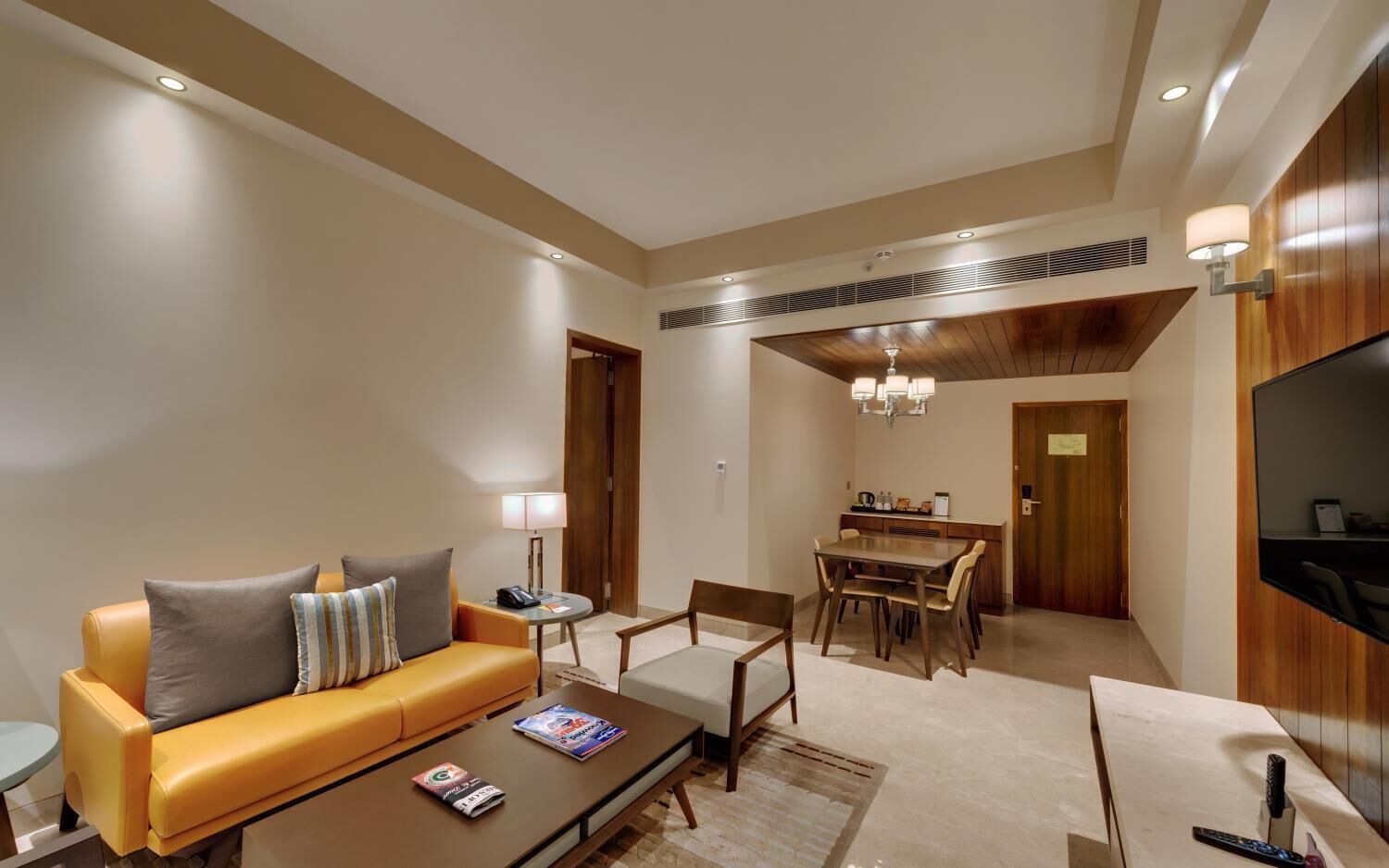 A hotel room with a sofa, coffee table, dining table, chairs, a TV, and a kitchen area.