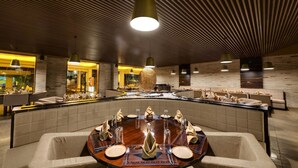 Un restaurant moderne doté d’une grande table ronde au centre, dressée pour un repas, et entouré d’espaces de détente au design contemporain et à l’éclairage chaleureux.
