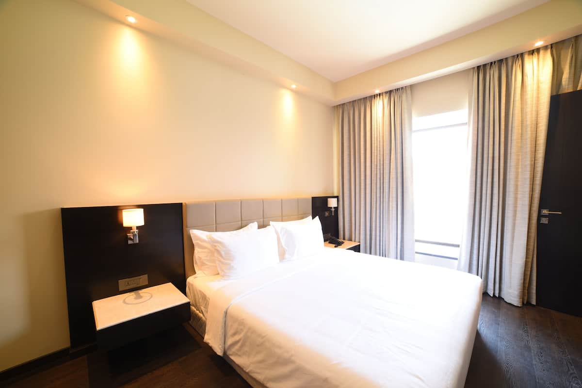 A hotel room with a large bed, a bedside table with a lamp, a nightstand, and a window with curtains.